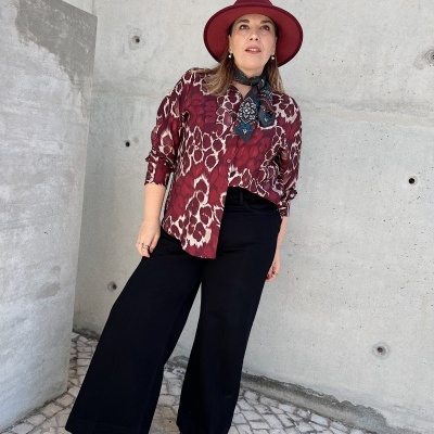 Mulher vestida com blusa vermelha estampada, calças pretas largas e chapéu vermelho em fundo de parede de betão.