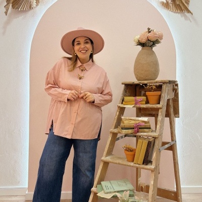 Mulher vestida com camisa rosa, calças de ganga e chapéu rosa junto a uma escada de madeira com livros e plantas.