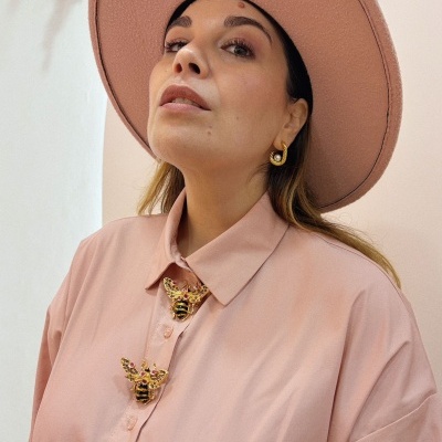 Mulher com camisa rosa claro com botões abelha dourada, chapéu rosa e brinco dourado com pérola