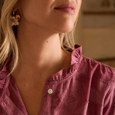 Blusa rosa texturizada com padrão floral e botões brancos, usada por mulher com brincos dourados e flor branca