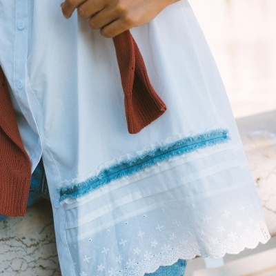 Camisola branca detalhada com renda e fita azul na bainha, calças de ganga e casaco castanho.