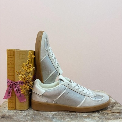 Sapatilhas prateadas com detalhes brancos e sola castanha claras em cima de uma mesa de madeira com livros e flores secas.