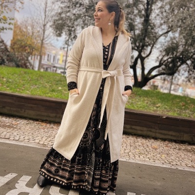 Mulher com casaco bege e vestido preto com padrões dourados em ambiente exterior