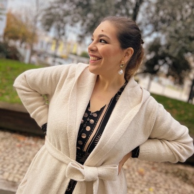 Mulher sorridente com casaco bege de malha com cinto, em ambiente exterior com árvores.