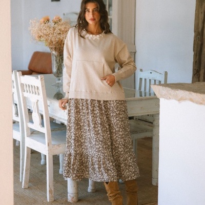 Mulher com camisola bege e saia floral castanha numa sala com mesa e cadeiras brancas.