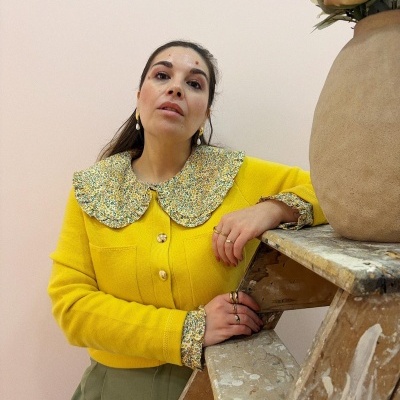 Mulher com camisola amarela com gola e punhos floridos encostada a escada com vaso e flores