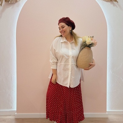 Mulher com boina vermelha, camisa branca, saia vermelha e sapatilhas segurando vaso com flores contra parede rosa com arco decorado.