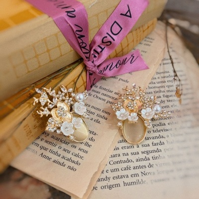 brincos dourados com flores brancas sobre páginas de livro e caixas com fita rosa