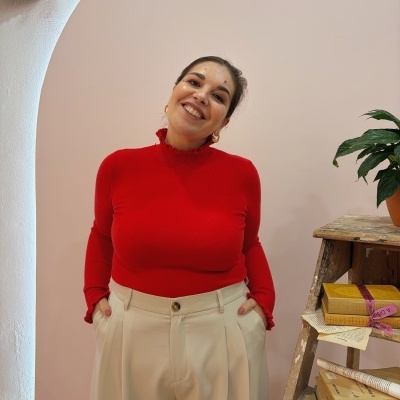 Mulher com camisola vermelha de manga comprida e calças bege junto a estante com livros e planta