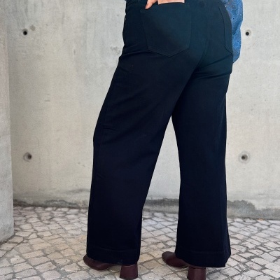 Calças pretas largas com camisa azul bordada e sapatos castanhos, cenário piso pedra e parede cimento.