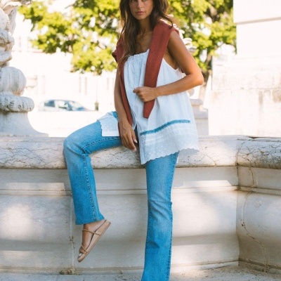 Mulher com blusa branca sem mangas, calças de ganga e casaco castanho ao ombro numa estrutura de pedra exterior.