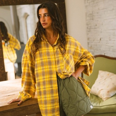 Mulher com blusa xadrez amarela e calções verdes acolchoados junto a mesa de madeira em sala com decoração rústica