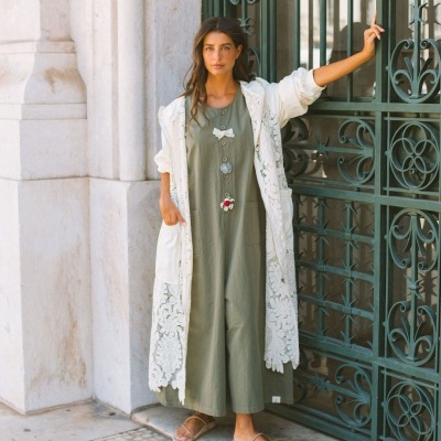 Mulher com vestido caqui e casaco rendado branco encostada em grade metálica verde