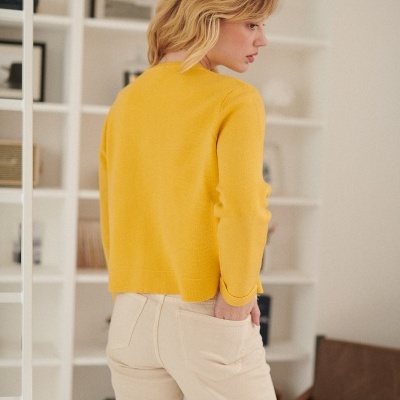 Mulher de costas com camisa amarela e calças bege numa sala com estantes brancas