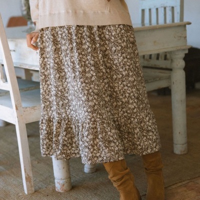 Saia midi floral castanha e bege com botins castanhos e casaco bege, ambiente interior com mobiliário rústico.