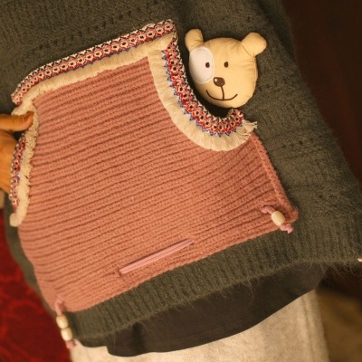 Mala rosa com figura de urso e bordas decorativas sobre fundo verde tricotado.