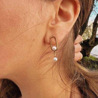 Brinco dourado com pérolas brancas usado numa orelha de mulher com cabelo castanho claro.