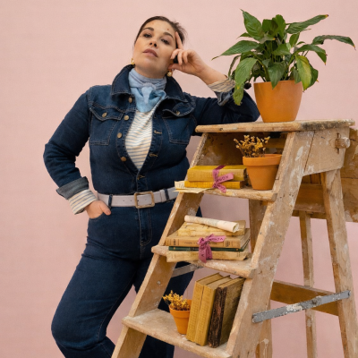 Mulher com macacão jeans ao lado de escada de madeira com livros e vasos, parede rosa ao fundo.