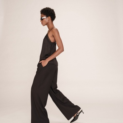 Mulher vestindo blusa preta de alças finas, calças largas pretas e sapatos de tacão, com óculos escuros em fundo claro.