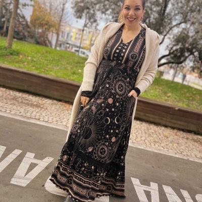 Mulher com vestido longo preto com padrão astronómico, casaco bege comprido e botins pretos num parque.