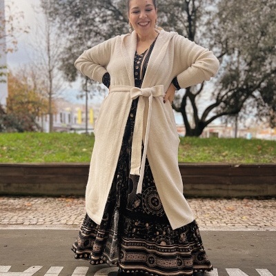 Mulher sorridente com casaco creme e vestido preto com padrão celestial, ao ar livre