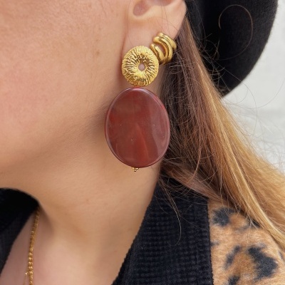 Brinco pendente dourado com peça oval vermelha castanha numa orelha, ao lado de casaco preto com padrão animal print e chapéu preto.