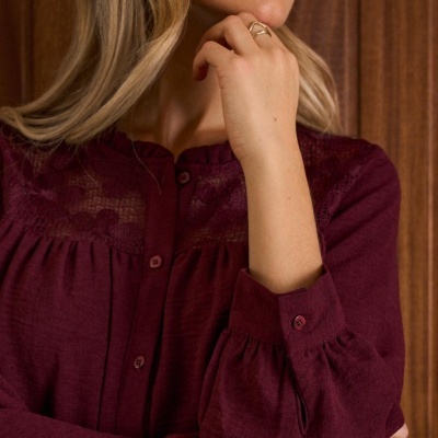 Blusa feminina vinho com renda e botões, vista até ao pescoço e braço dobrado com anel dourado.