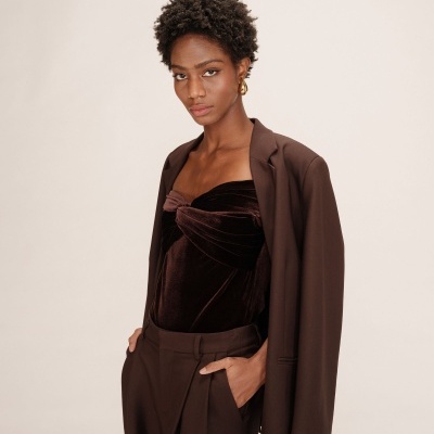 Mulher a usar conjunto castanho escuro com top de veludo e casaco por cima, fundo neutro claro.