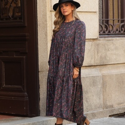 Mulher com vestido comprido estampado e chapéu preto numa rua com parede de pedra.