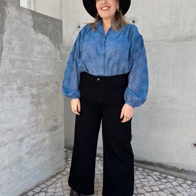 Mulher com camisa azul, calças pretas largas, chapéu preto e sapatos castanhos escuros em piso de calçada portuguesa