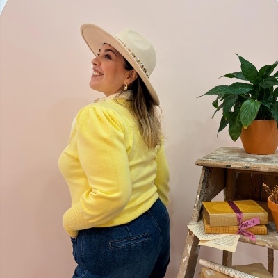 Mulher de perfil com camisola amarela, jeans e chapéu, ao lado de estante com livros e planta.