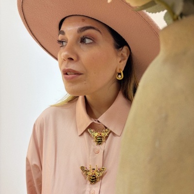 Mulher com camiseiro rosa claro e broches de abelha dourados veste chapéu rosa e brincos dourados com pérola