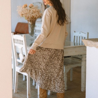 Mulher de costas com camisola bege, saia floral e botas castanhas em ambiente rústico interior