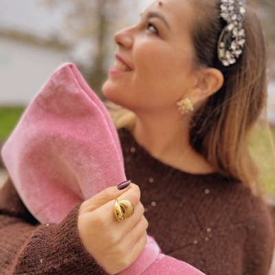 Mulher sorridente com camisola castanha, anel dourado, tiara de cristais e casaco rosa macio ao ar livre