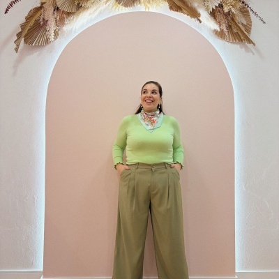 Mulher com roupa verde e lenço floral em frente a parede cor de rosa com arco iluminado e flores secas no topo