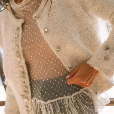 Cardigan bege com botões decorativos sobre blusa transparente com padrão de corações e folho.