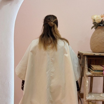 Pessoa de costas com casaco bege junto a escada de madeira com livros e jarro de flores