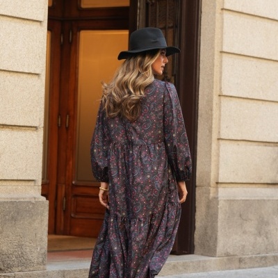 Mulher de costas com vestido longo padrão floral, botas de salto castanhas e chapéu preto numa rua com porta e parede de pedra.