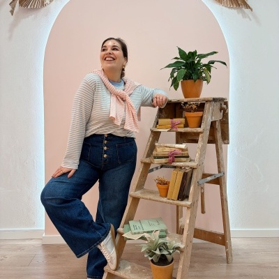 Escada de madeira com plantas, livros e mulher sorridente ao lado em fundo rosa claro.