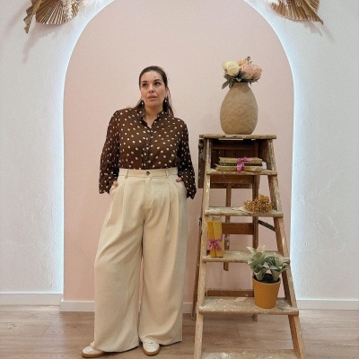 Mulher com blusa castanha de bolinhas brancas e calças largas creme, ao lado de escada de madeira com decoração