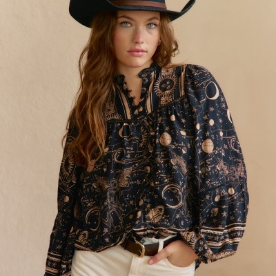Blusa feminina preta com estampas astrocelestes e detalhes em babado, combinada com calças claras e chapéu preto.