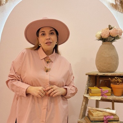 Mulher com camisa rosa e chapéu rosa em ambiente decorado com livros e flores.