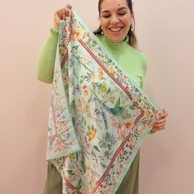 Lenço quadrado floral colorido em fundo verde claro segurado por mulher com blusa verde claro.