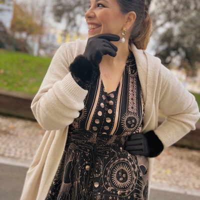 Mulher com vestido astrológico preto e dourado, casaco creme e luvas pretas ao ar livre