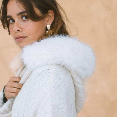 Mulher com casaco branco de malha com capuz de pelo branco e brincos dourados