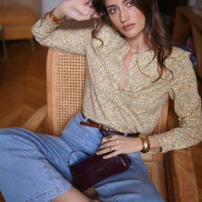 Mulher sentada numa cadeira de madeira com blusa floral, calças de ganga azuis e acessórios dourados