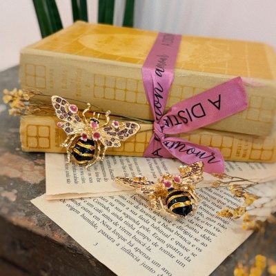 Dois broches de abelha dourados, pretos e rosas sobre papel e livros amarelos com fita rosa.