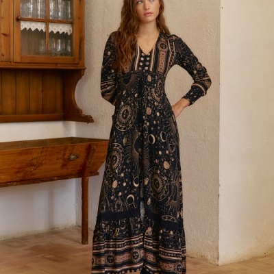 Vestido comprido preto com padrões astrológicos bege e botas pretas, em ambiente interior rústico.
