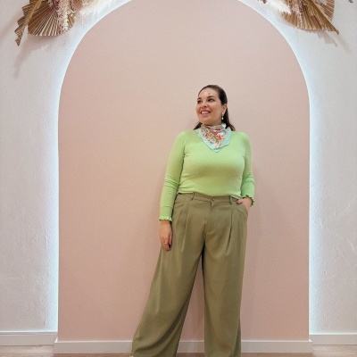 Mulher com camisola verde clara, lenço floral, calças largas verde cáqui e sapatilhas verdes em fundo rosa com arco decorado.