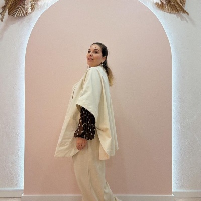 Mulher com capa bege e blusa castanha com bolas, em parede rosa com arco decorativo